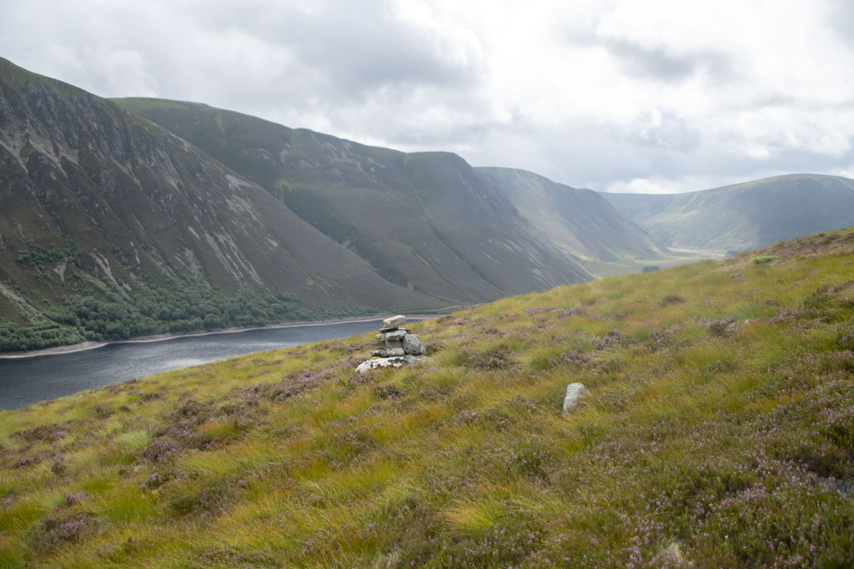 A small stone cairn stands on a grassy hillside covered in patches of purple heather, overlooking a dark loch nestled between steep, rolling mountain slopes under an overcast, cloudy sky.